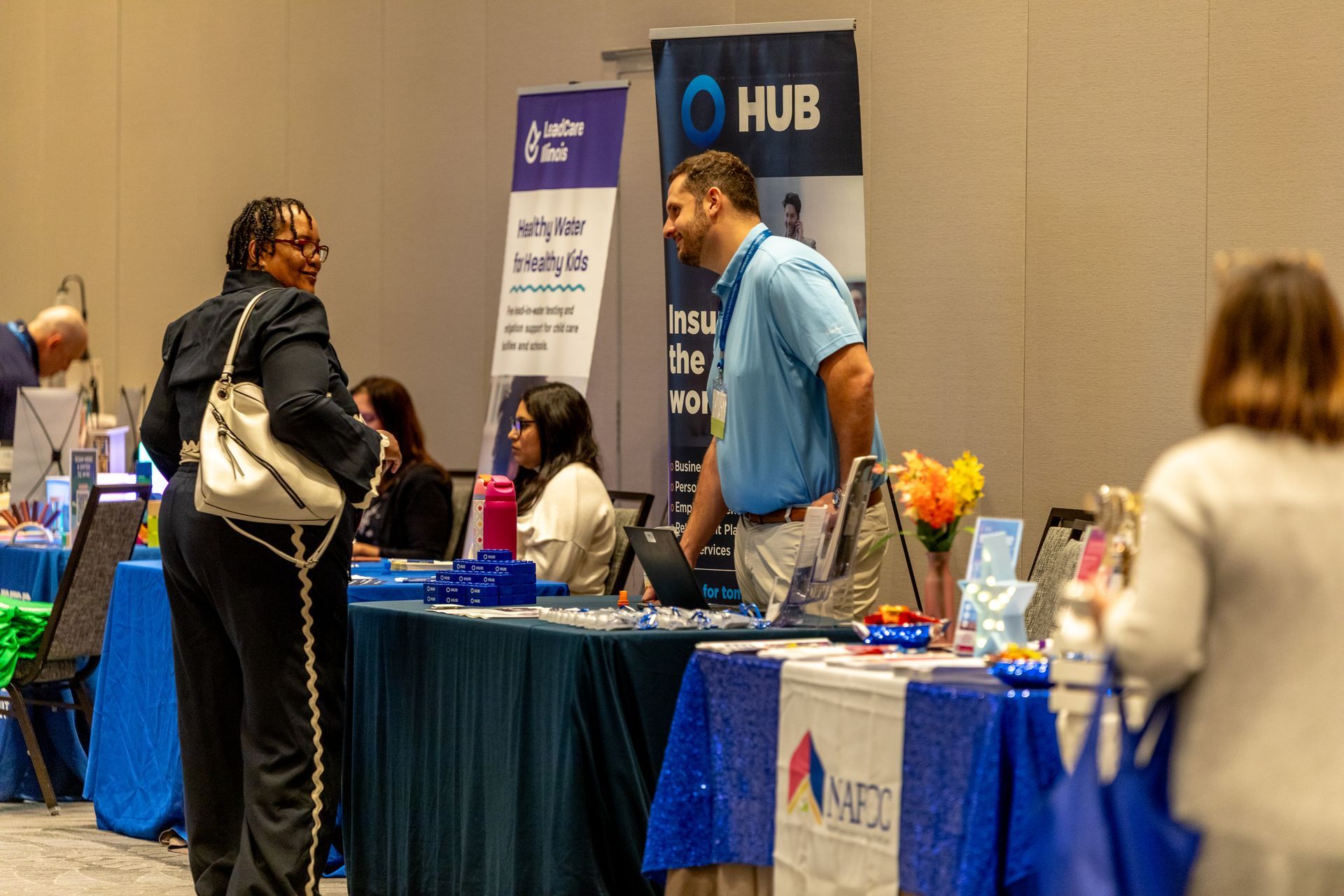 People talking at a booth with a blue tablecloth at a conference expo, with a “HUB” sign in the background