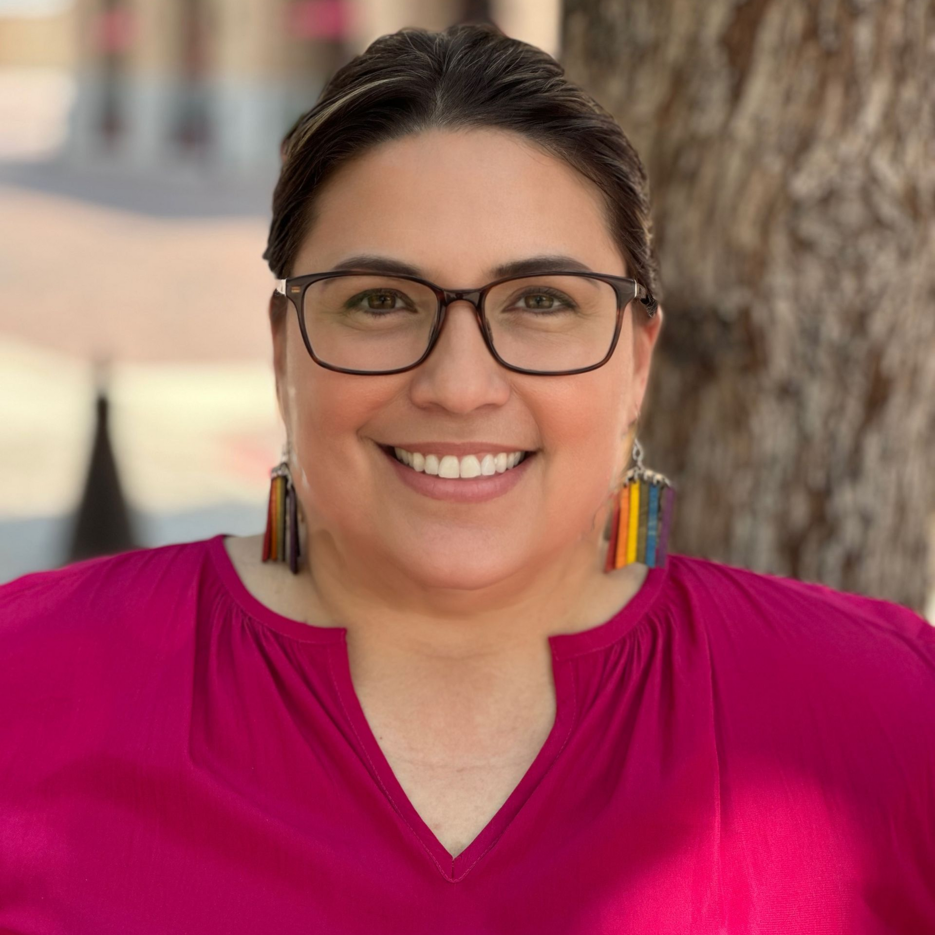Woman smiling, wearing glasses and colorful earrings, in front of a tree, wearing a pink blouse.