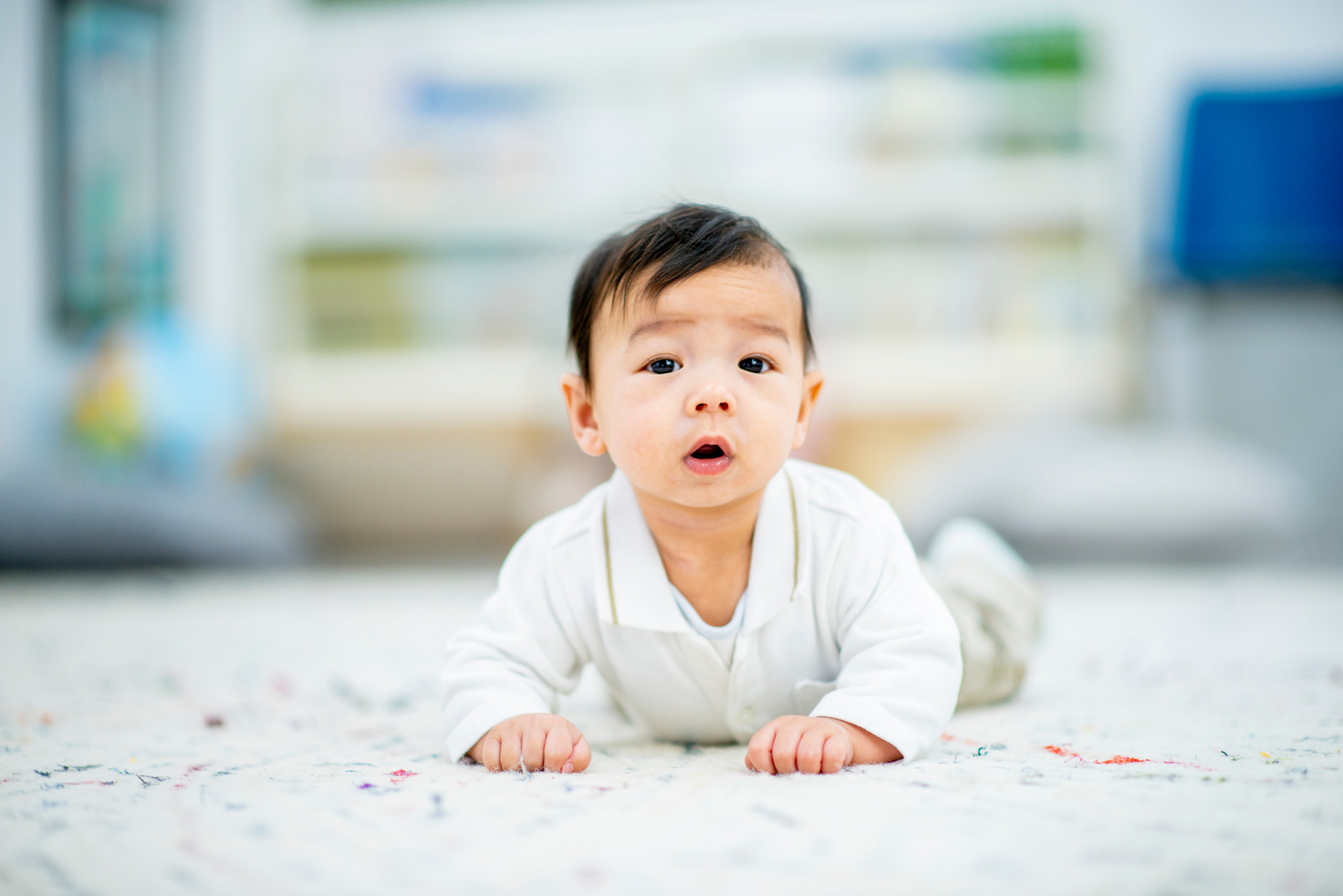 Baby in white outfit lying on a patterned floor, looking at the camera with mouth open.