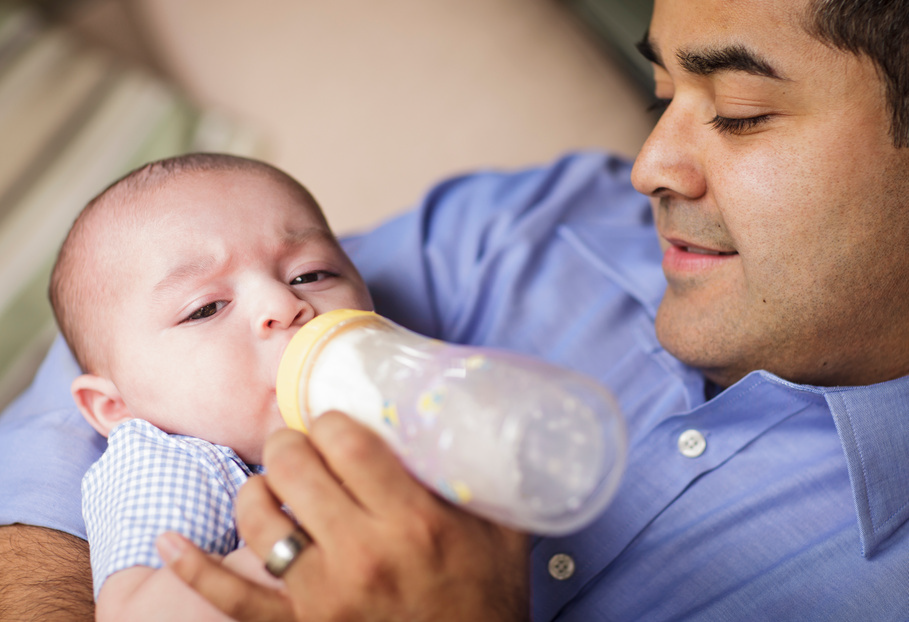 Man feeding a baby with a bottle, both in blue clothing, close-up indoors