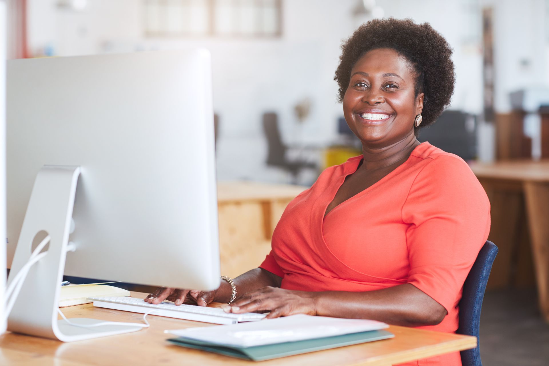 Smiling person in an orange shirt working at a desk with a computer in a bright office