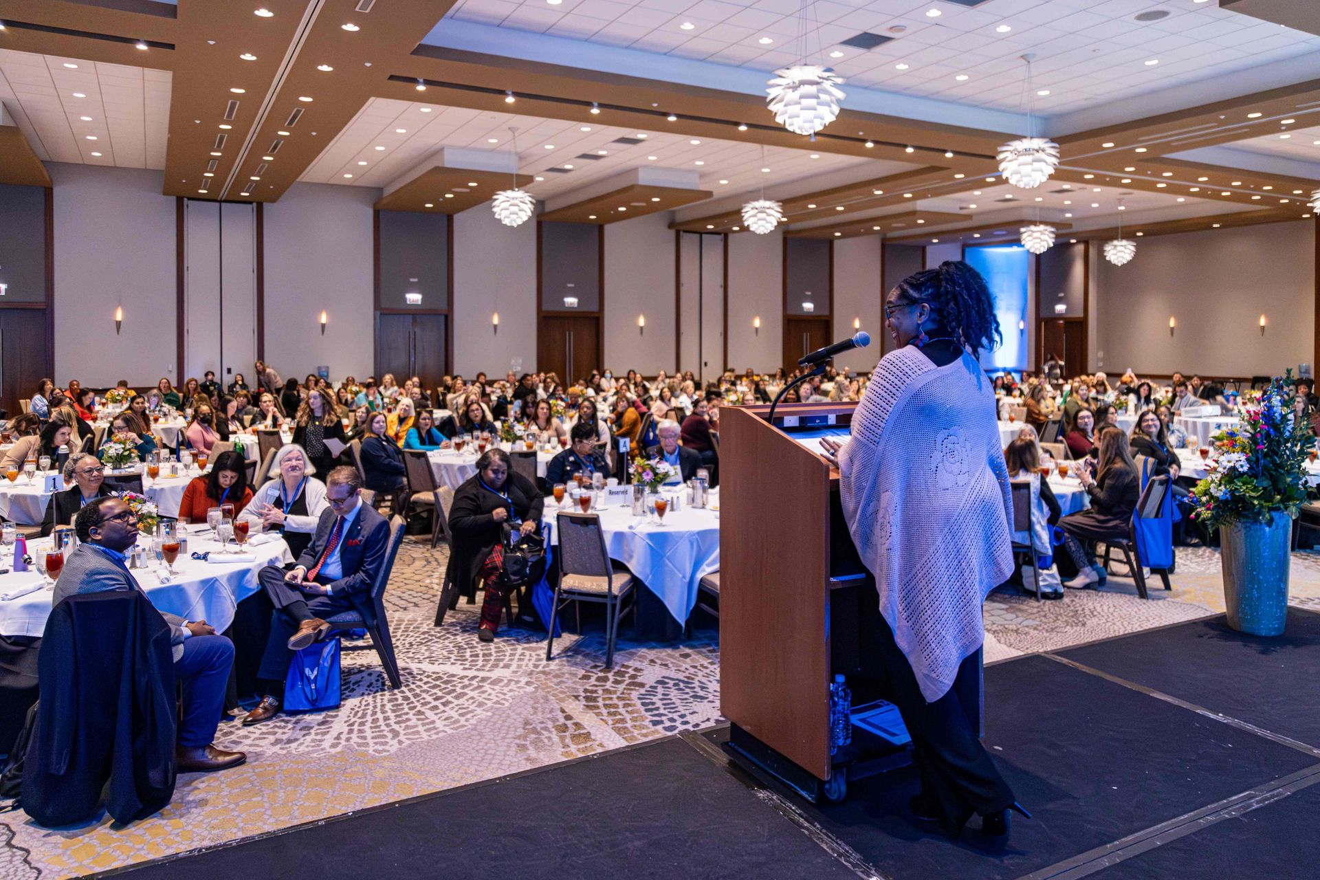 A woman speaking at a podium in a large banquet hall, addressing a diverse crowd seated at round tables.