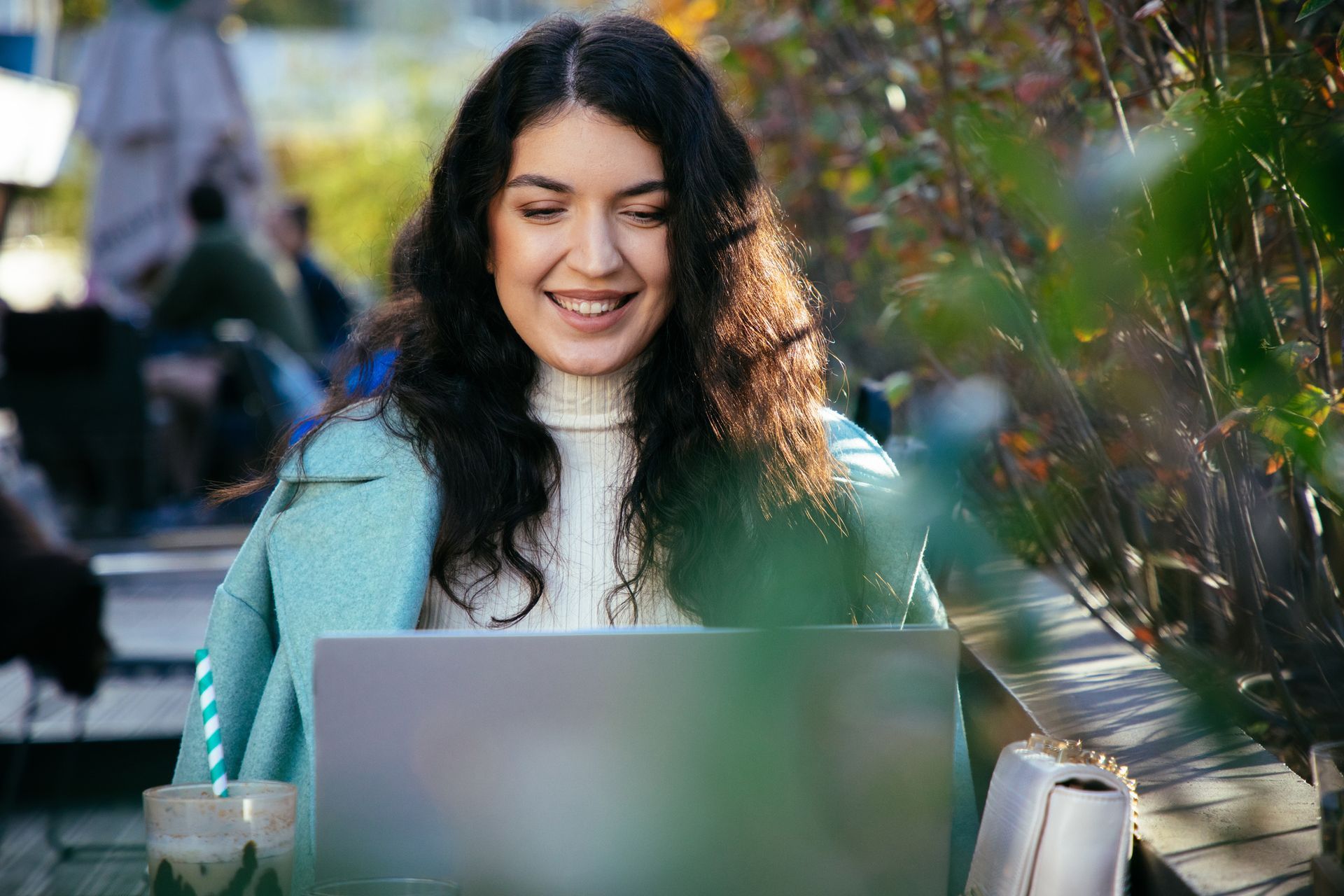 Smiling woman using a laptop at an outdoor café, with a light blue coat and soft bokeh background.