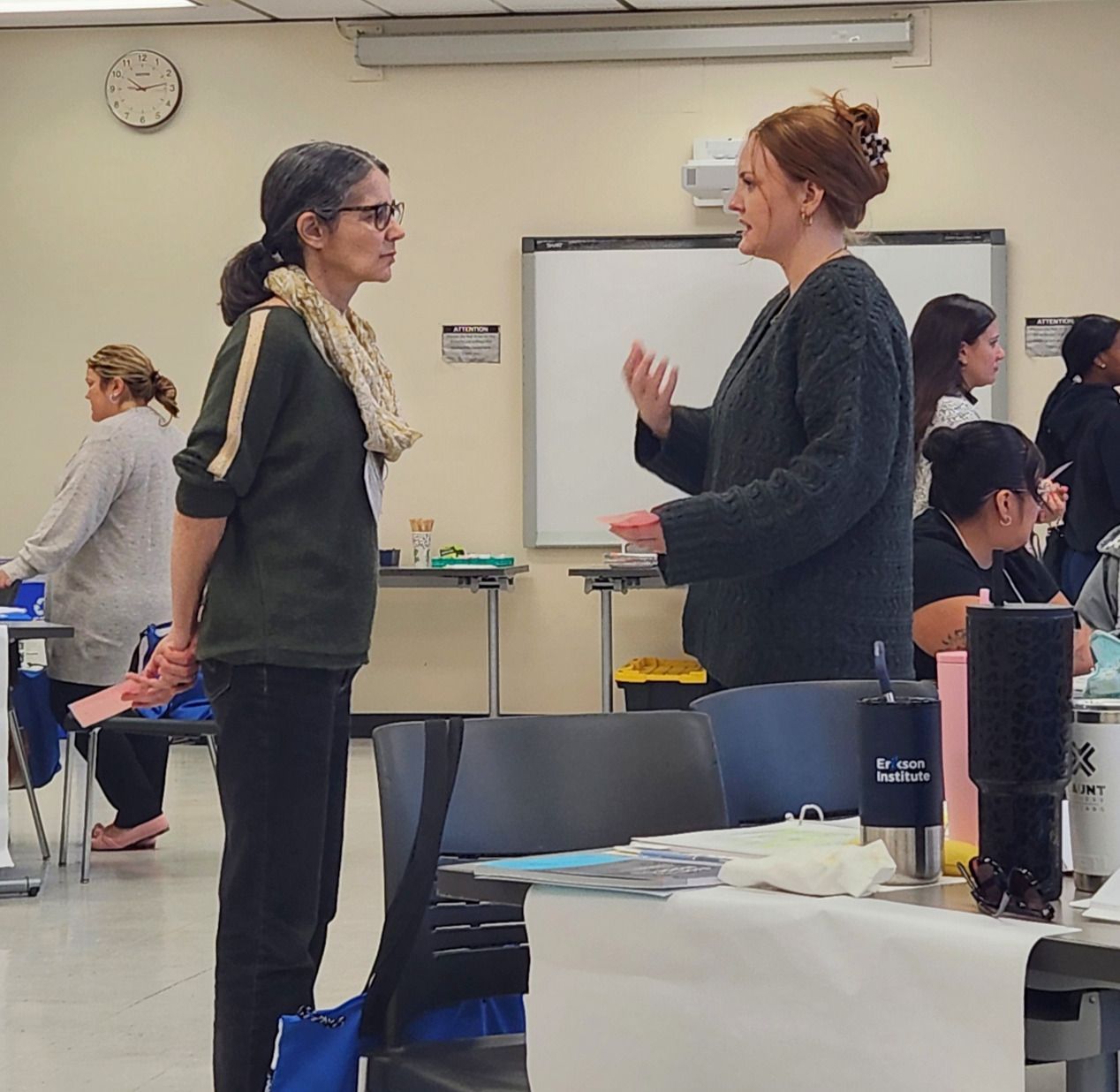 Two people talking in a classroom, with chairs, a whiteboard, and students in the background.