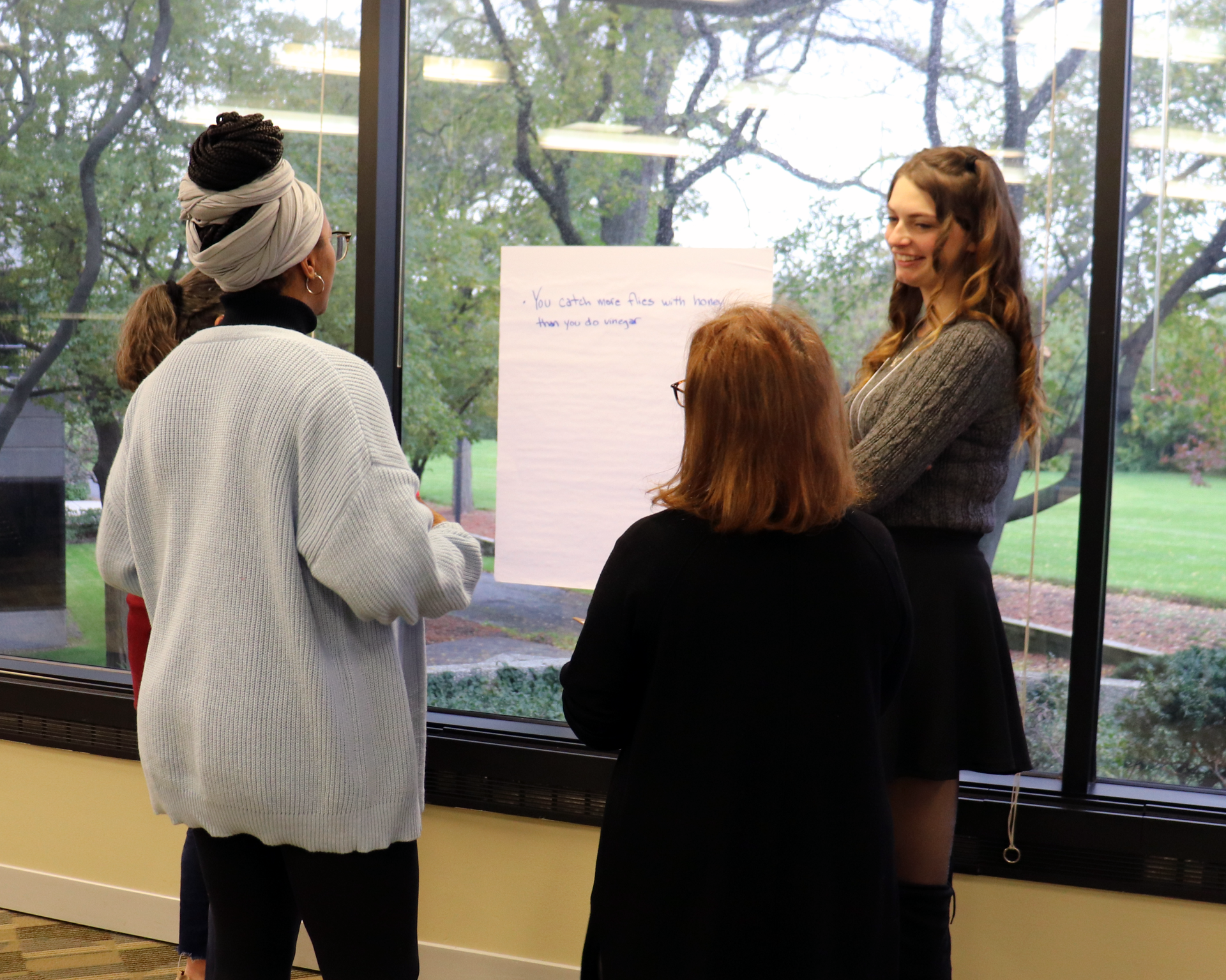 Three people stand by a window, discussing a poster board in a bright room.