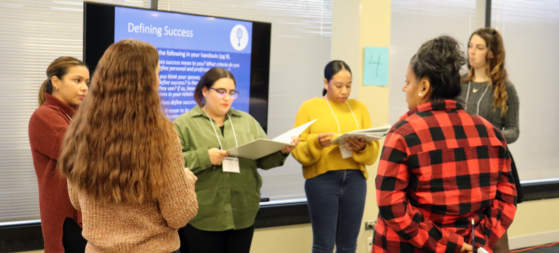 Group of people in a bright room, one presenting while others listen near a blue “Safety” sign.