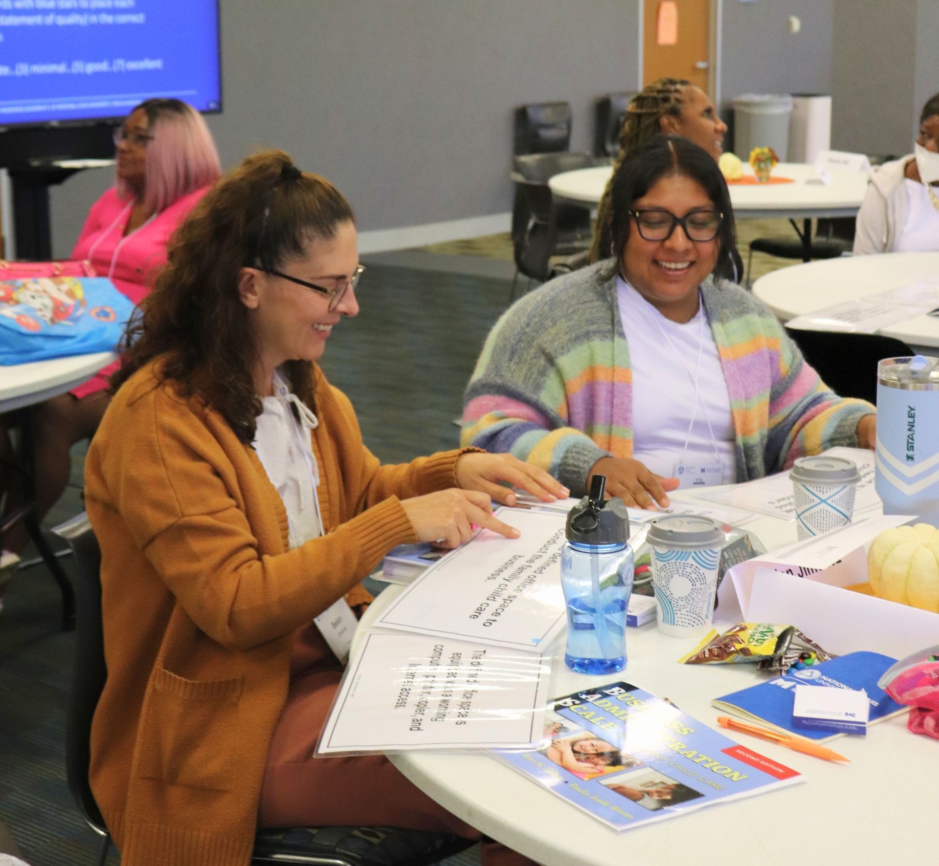 Two women smiling and working at a table with papers, snacks, and drinks in a conference room.