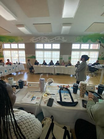 Group meeting in a bright classroom, people seated around tables while one person presents at the front.
