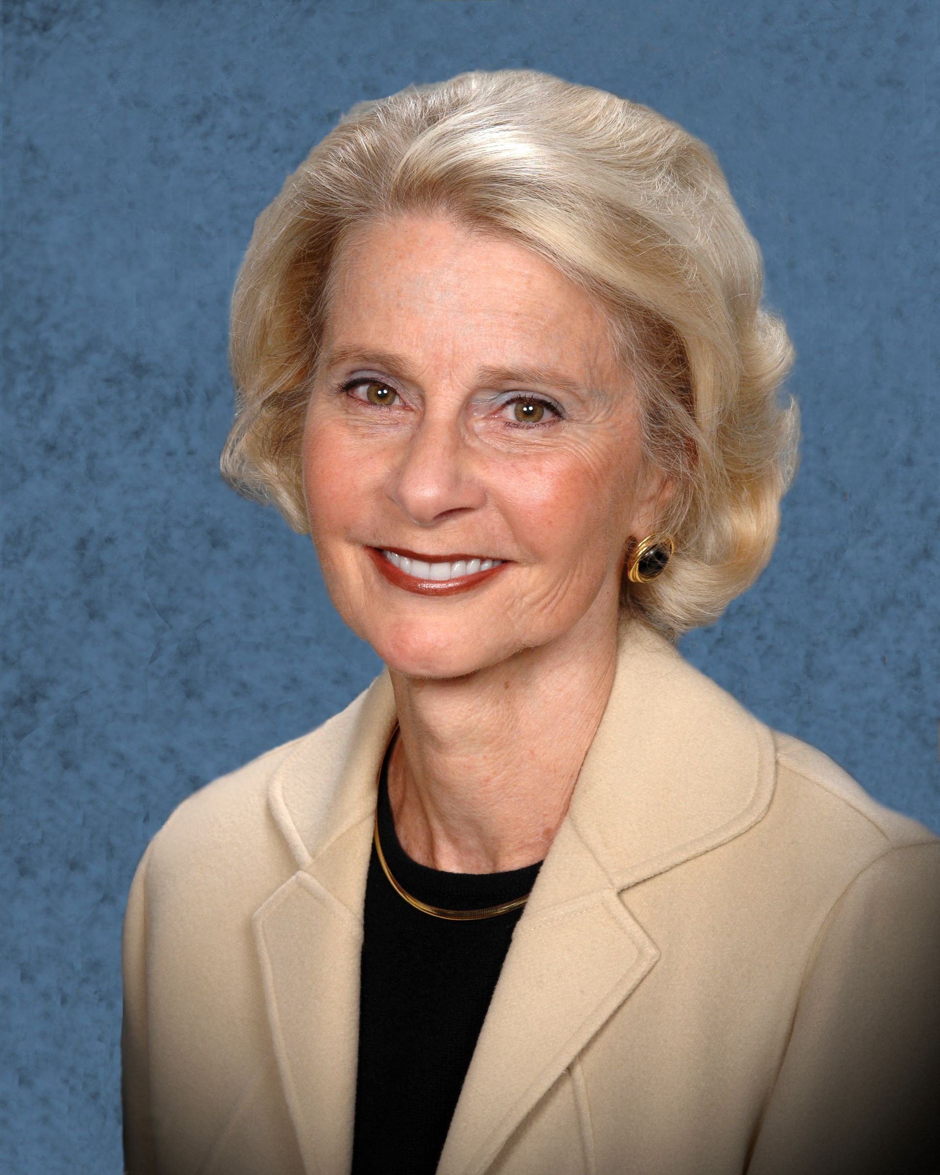 Woman with blonde hair wearing a beige blazer and black shirt, smiling against a blue background.