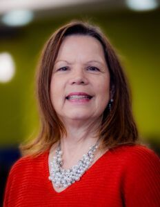 A woman wearing a red sweater and a pearl necklace is smiling for the camera.