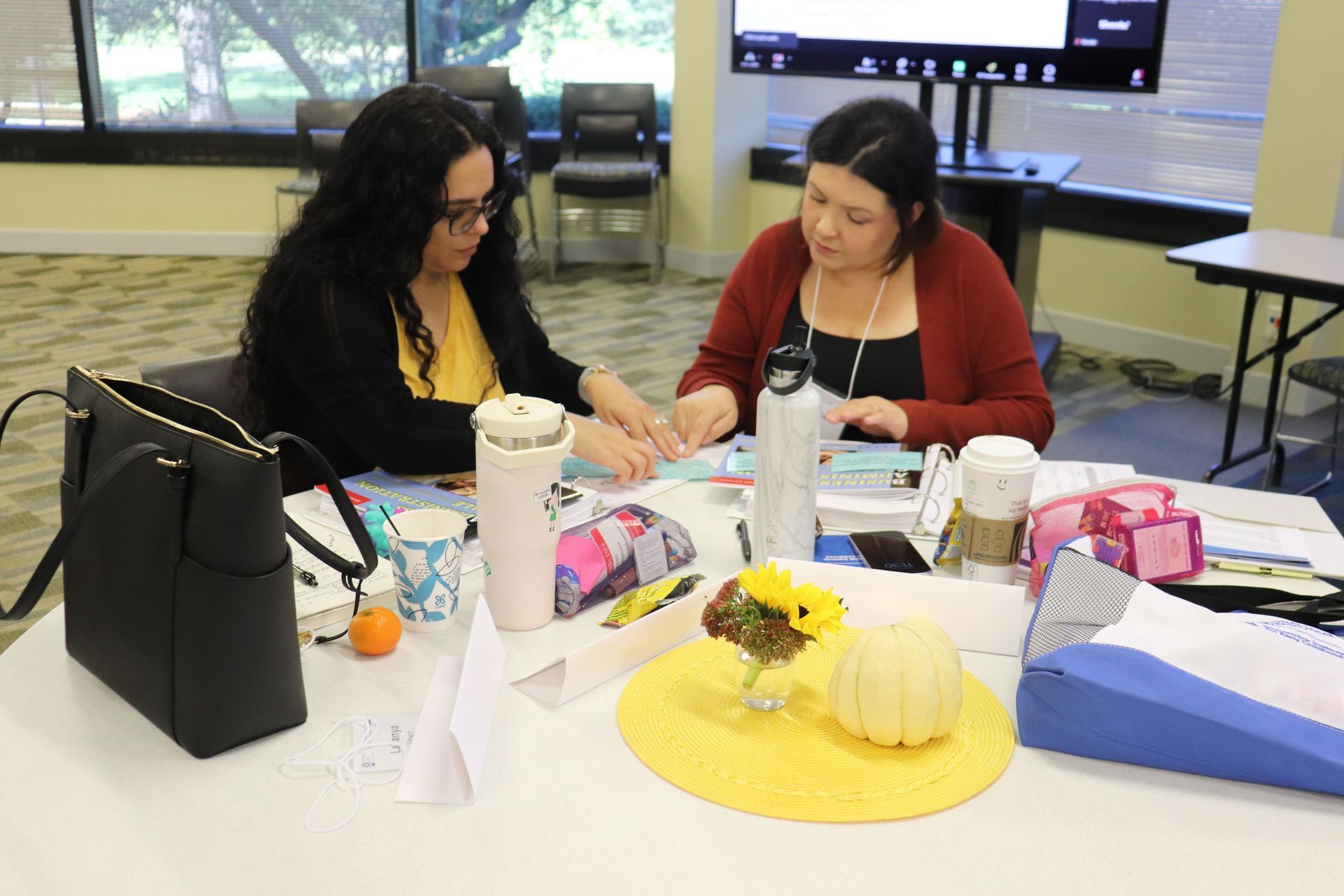 Two women collaborate at a table with papers, drinks, and craft supplies in a bright room.