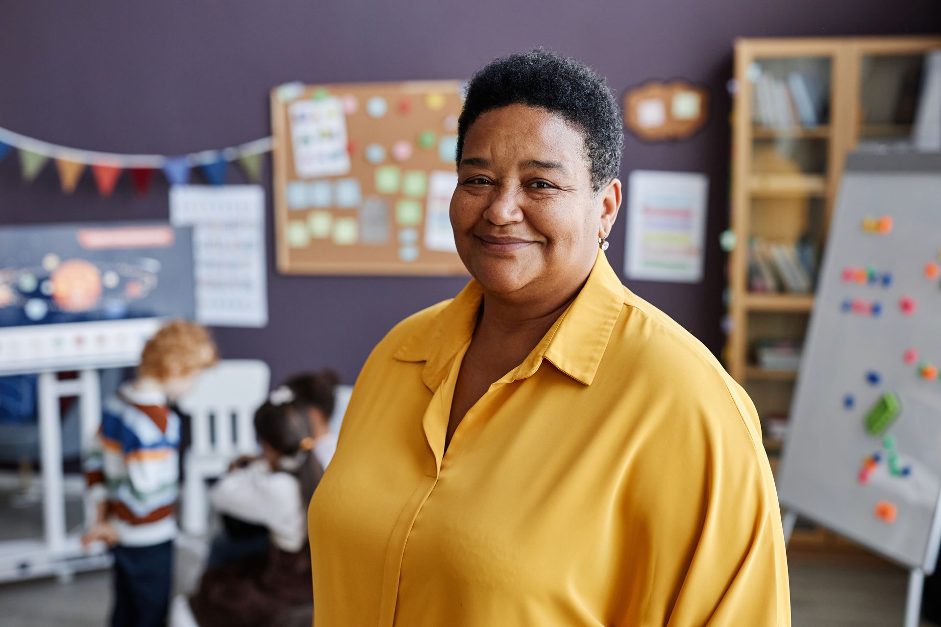 Teacher in a yellow shirt smiles in a classroom with children and colorful bulletin boards.