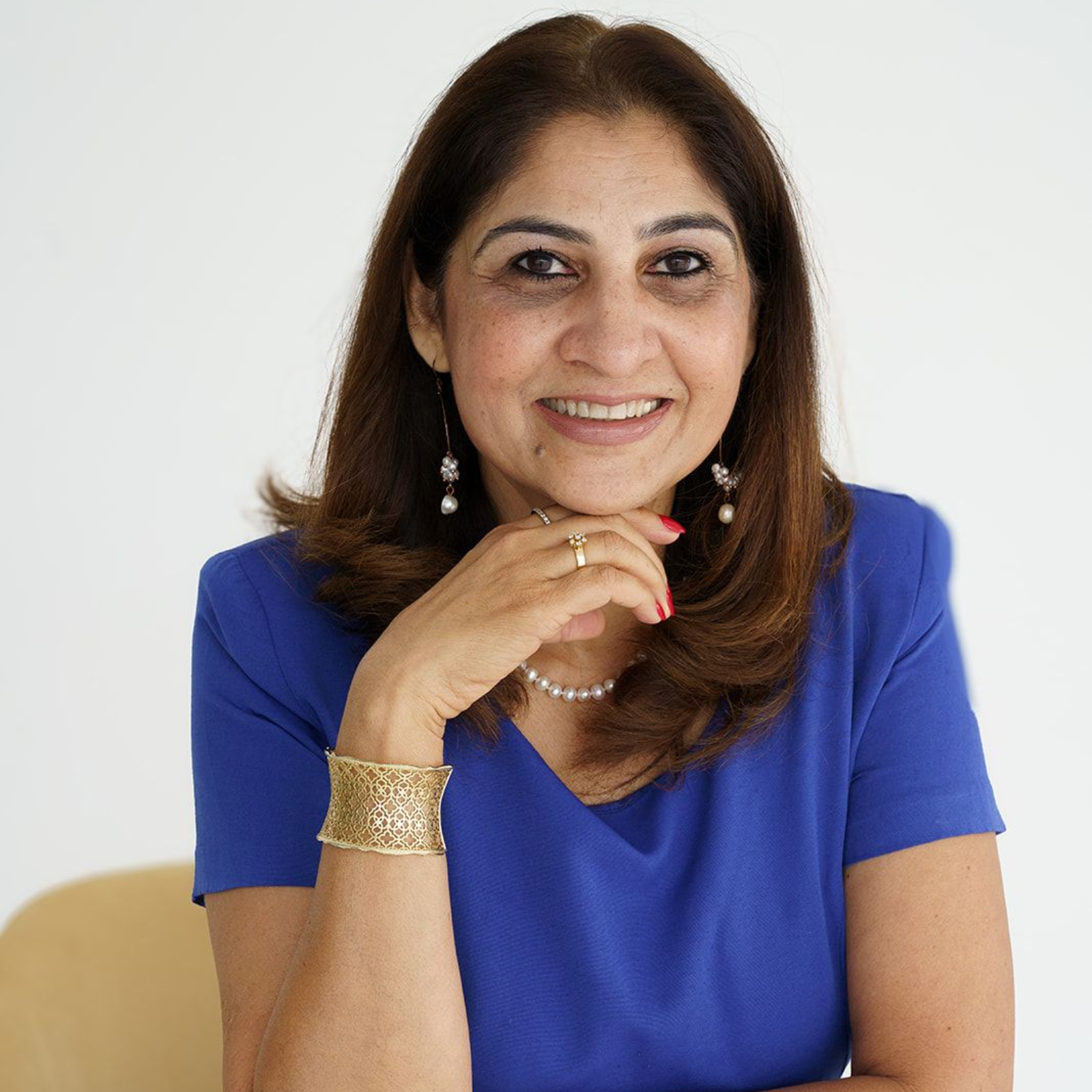 Woman smiling, wearing a blue top, gold bracelet, pearl necklace, and earrings, hand on chin.
