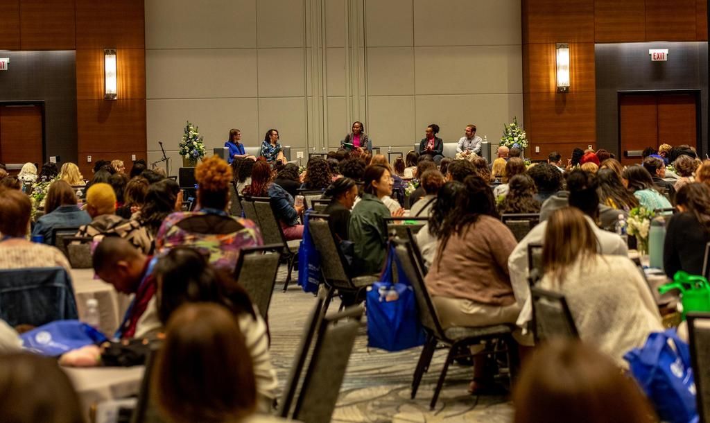Conference hall with many people seated facing a stage at the front, viewed from the back.