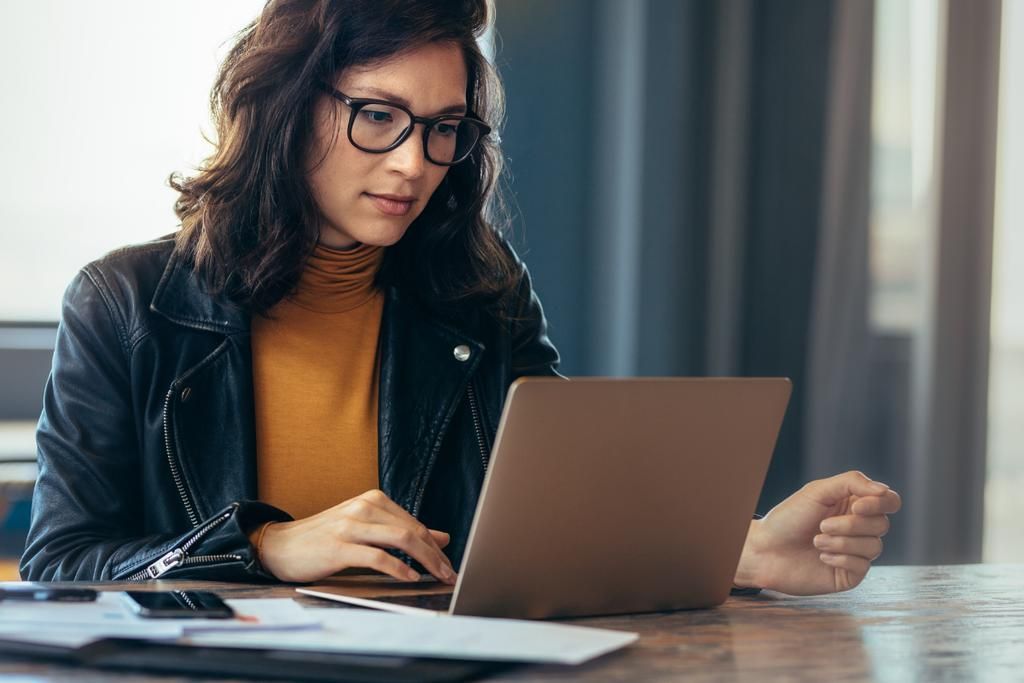 Person working on a laptop at a table, wearing glasses and a black jacket in a bright room