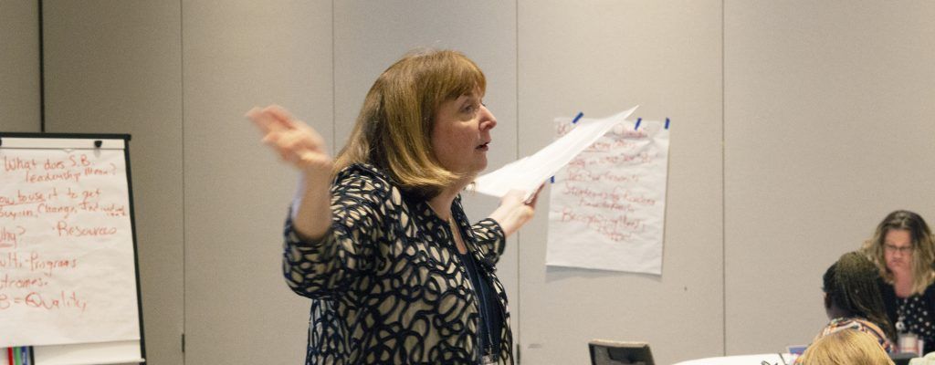 A woman is standing in front of a group of people holding a piece of paper.