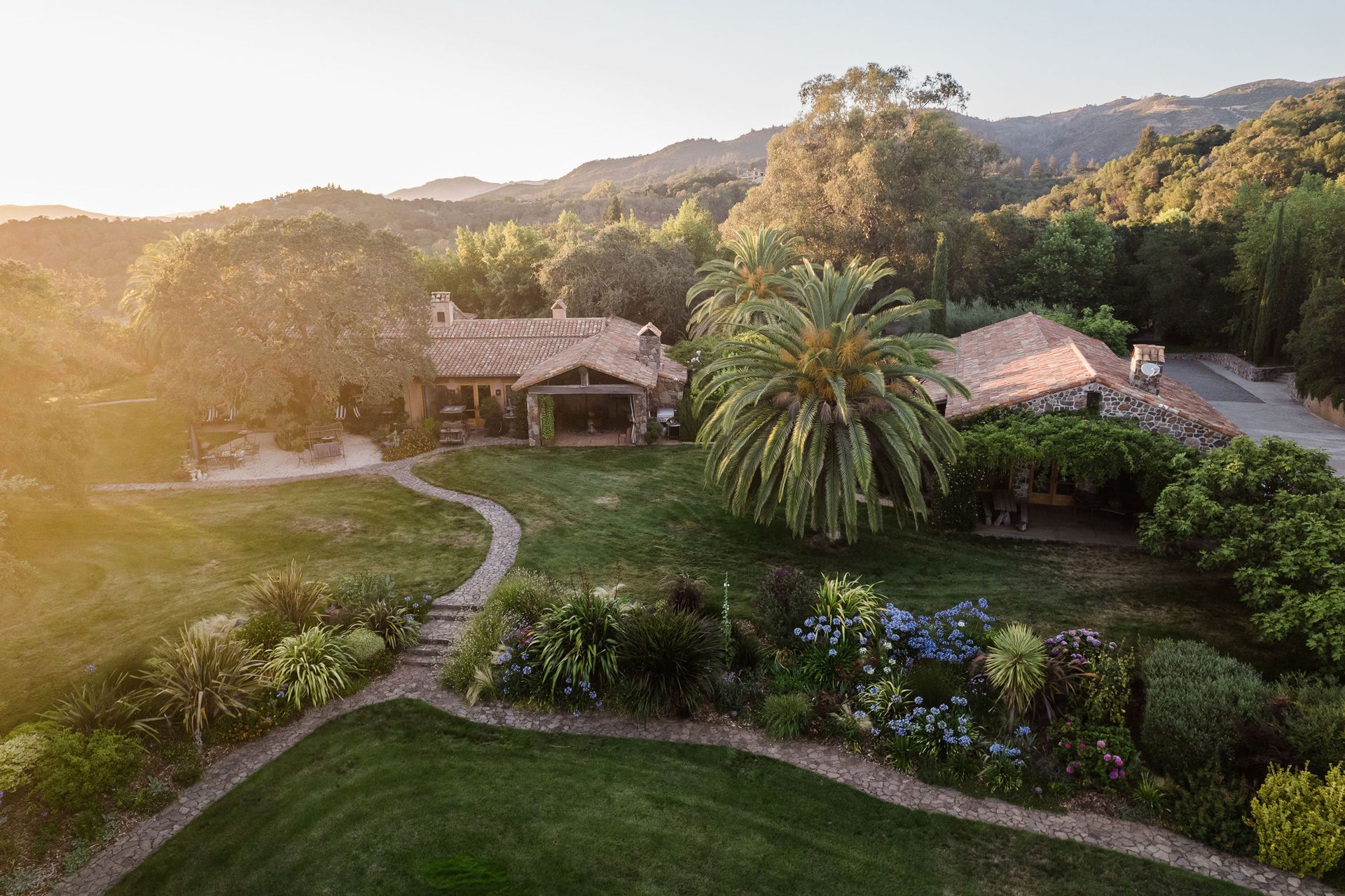 Sunlit hillside estate with stone cottages, palm tree, and winding garden paths among lush greenery