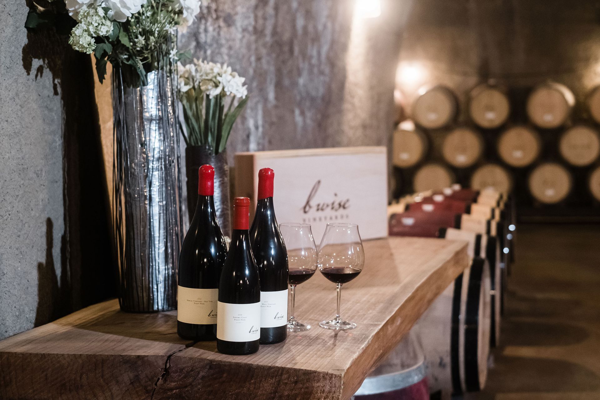 Wine bottles and glasses on a wooden table in a rustic tasting room with barrel decor.