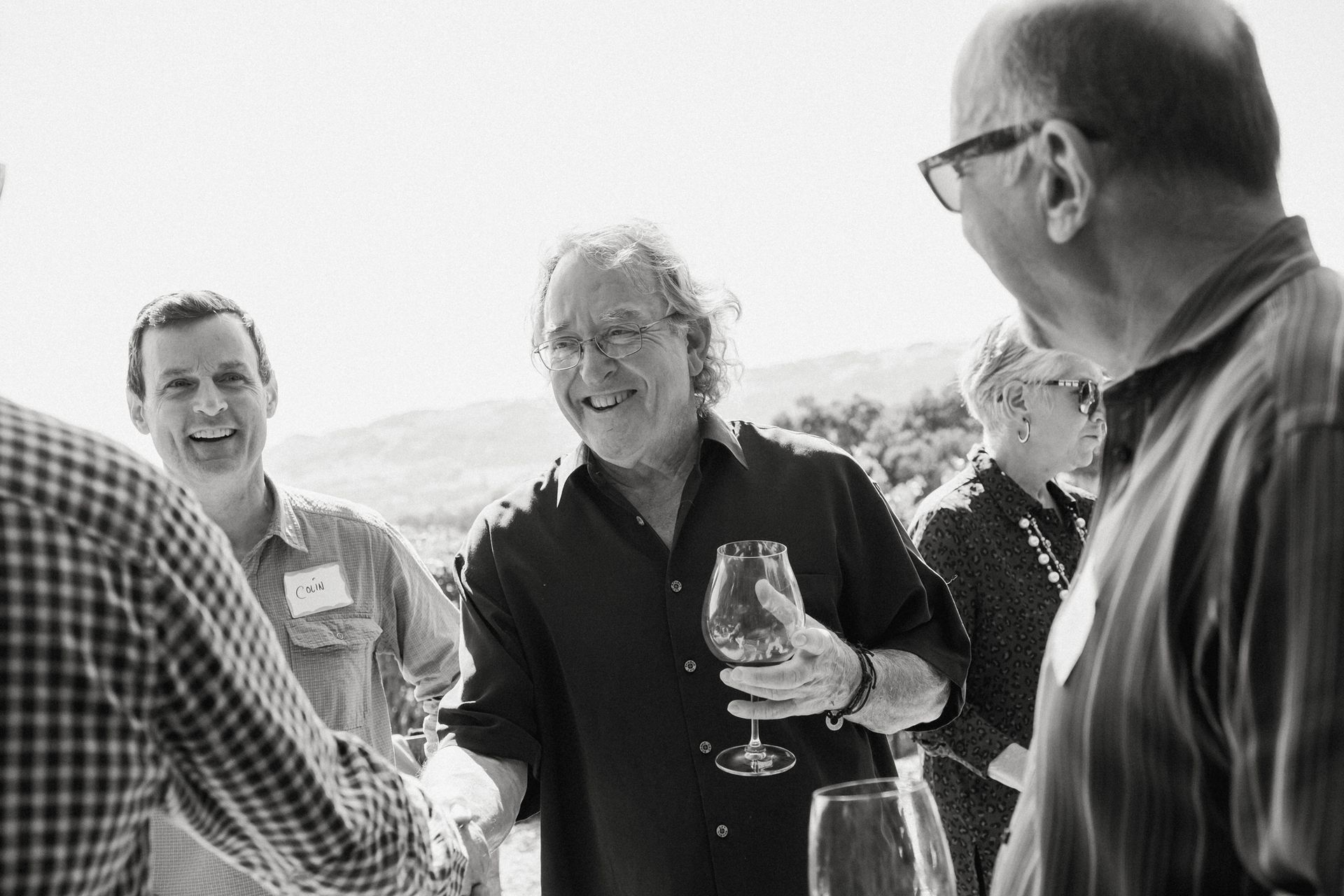 People chatting and laughing outdoors, one holding a wine glass, in a bright casual gathering
