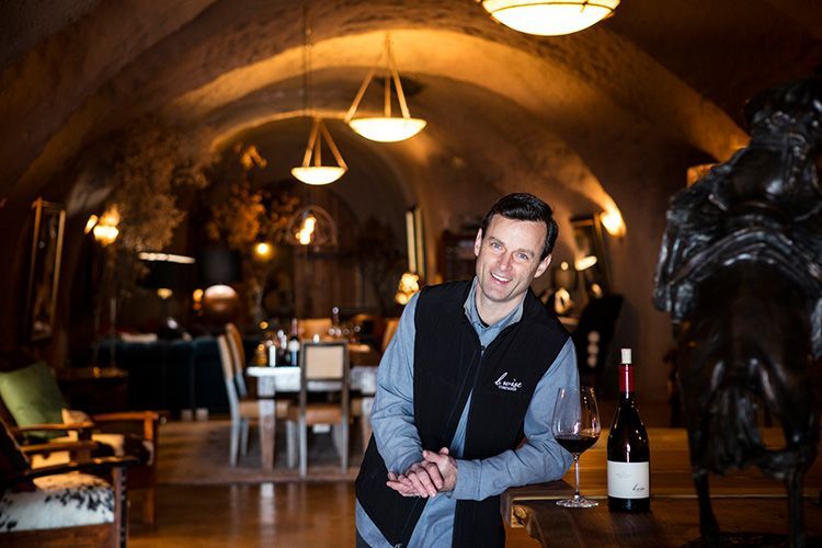 Smiling man in a dark vest stands in a warm, arched wine cellar with tables and bottles.