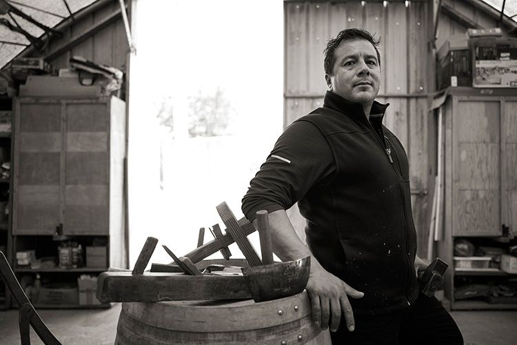 Man standing beside a barrel in a workshop, with tools and wooden structures in the background.
