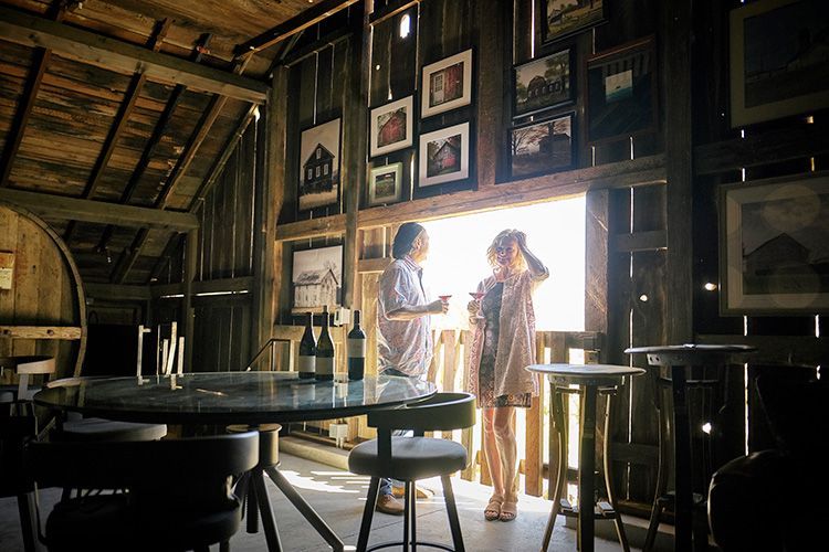 Two people standing in a sunlit rustic café doorway, with tables, stools, and framed photos on the walls.