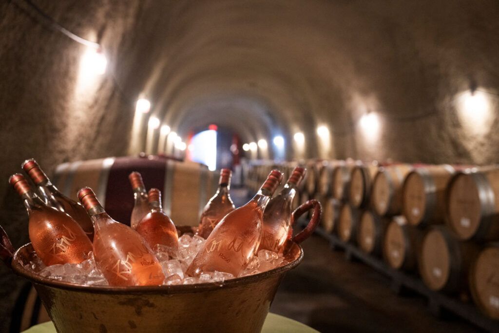 Copper bucket of chilled bottles in a wine cellar tunnel with stacked oak barrels and warm light