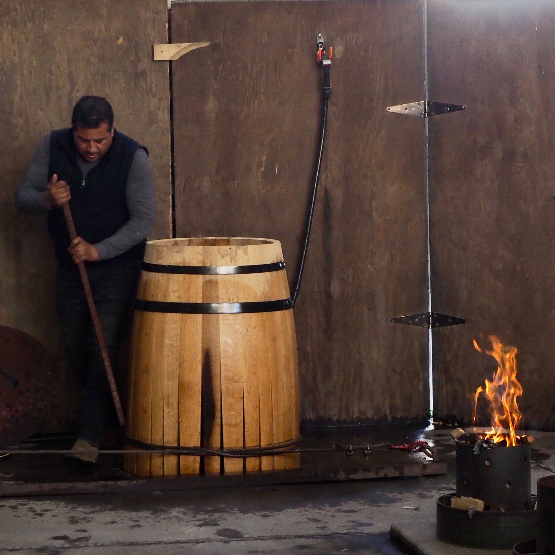 Worker stirs a wooden barrel beside a small flame in an industrial room with wet concrete walls.