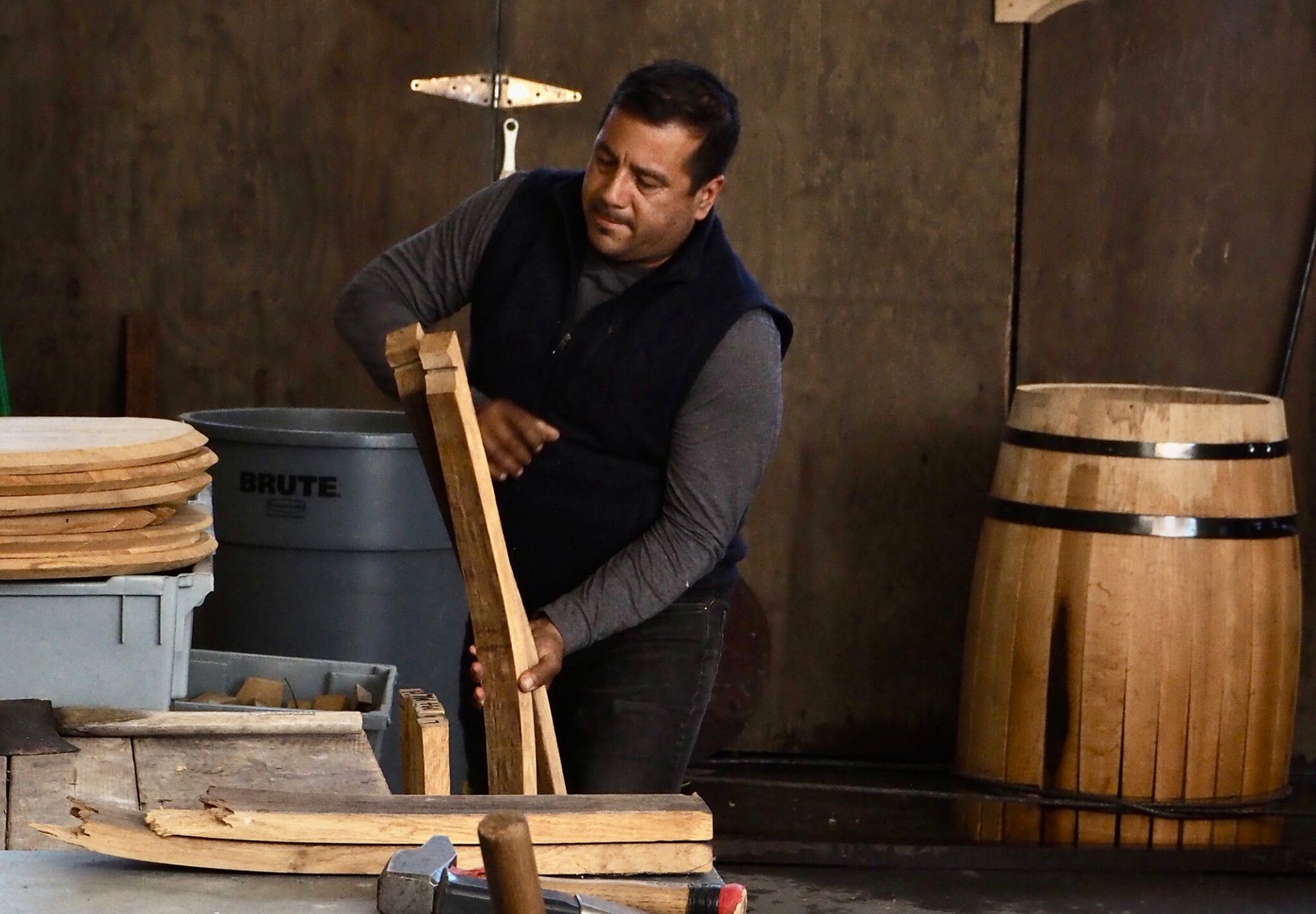 Man shaping wooden staves in a workshop, with barrels and timber nearby.