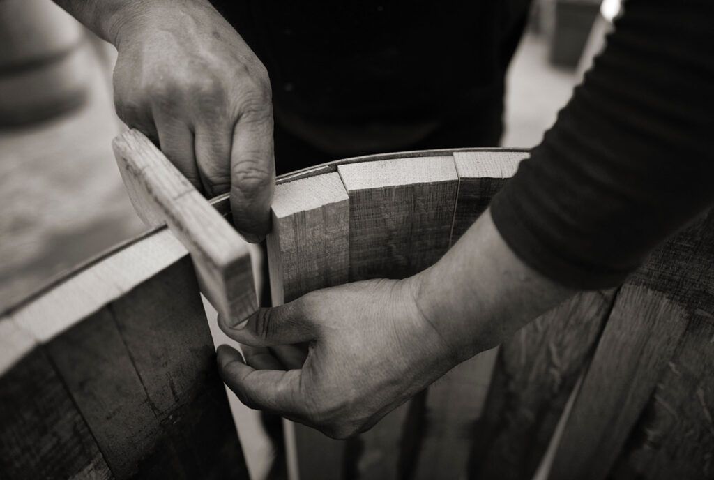 Hands weaving wood strips into a barrel-like basket in grayscale close-up