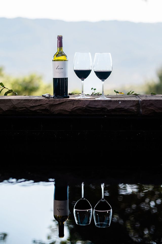Wine bottle and two glasses on a ledge, reflected in calm water with a misty background.