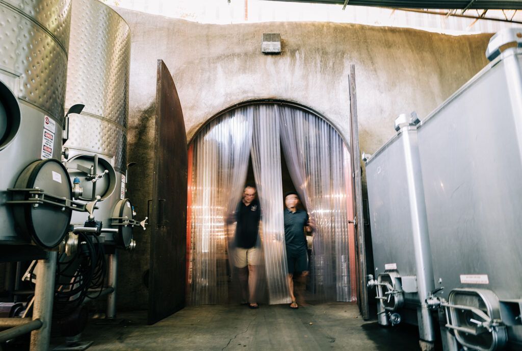 Two people standing in a winery cellar doorway between large stainless steel tanks