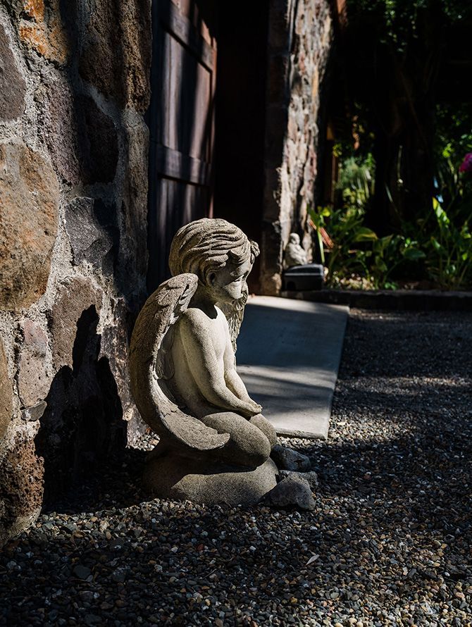 Stone cherub statue beside a wall in a sunlit garden path with gravel and greenery