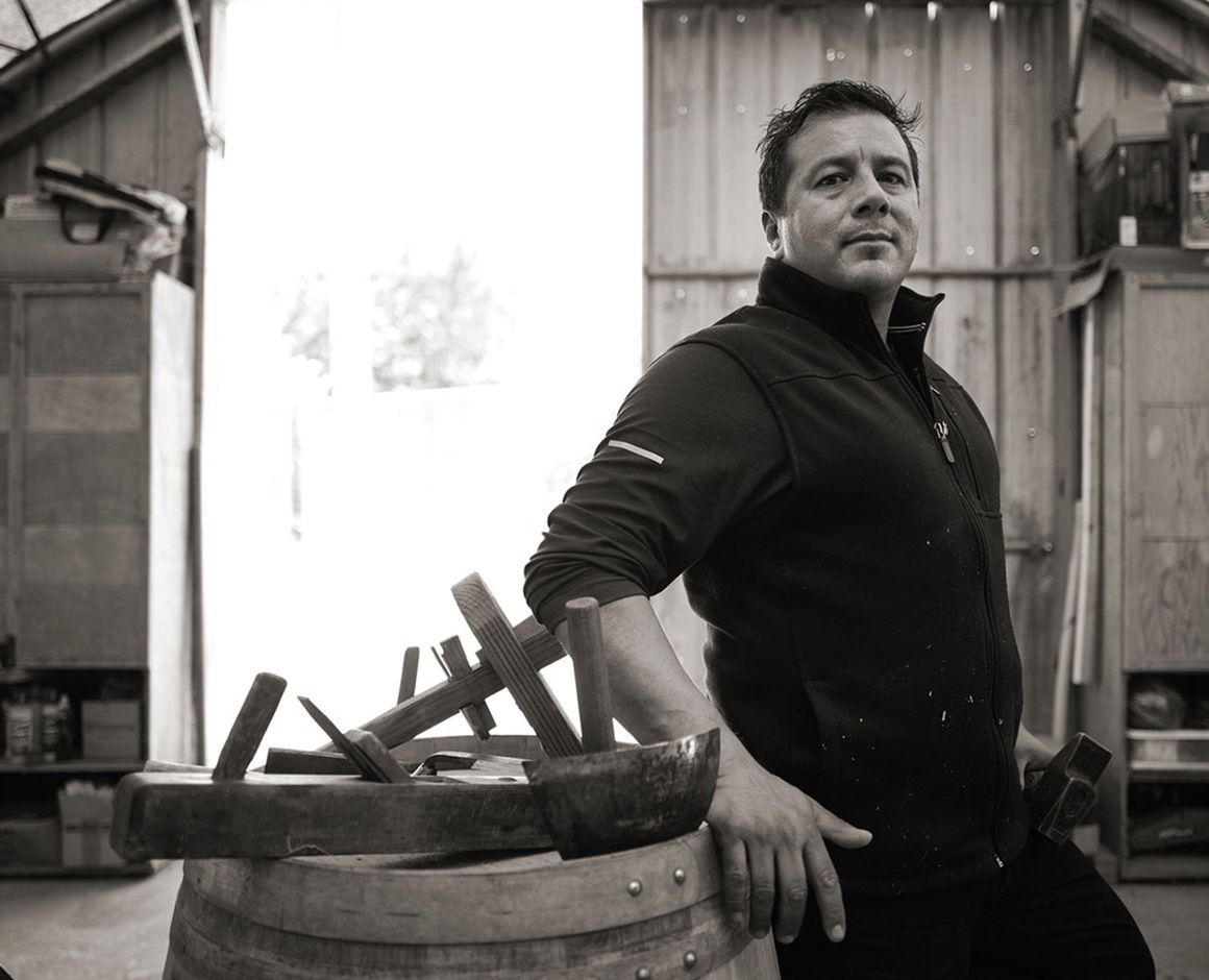 Man leaning on a barrel in a workshop, with tools and wooden planks nearby.