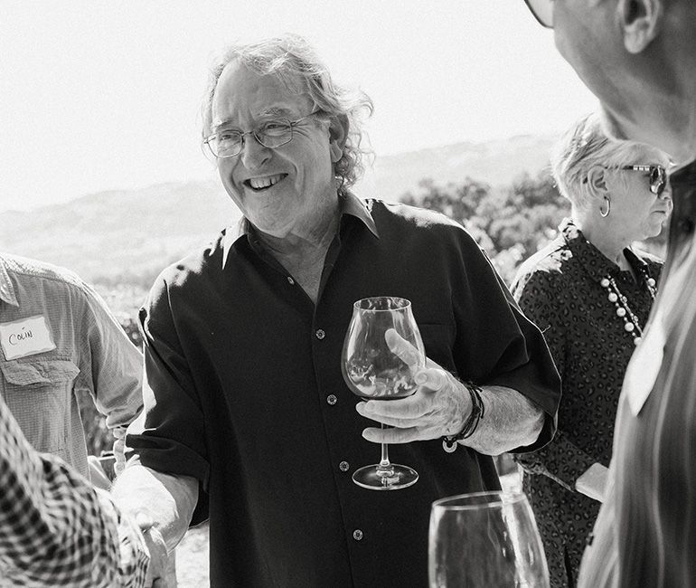 Smiling man in sunglasses holds a wine glass at an outdoor gathering, with others nearby.