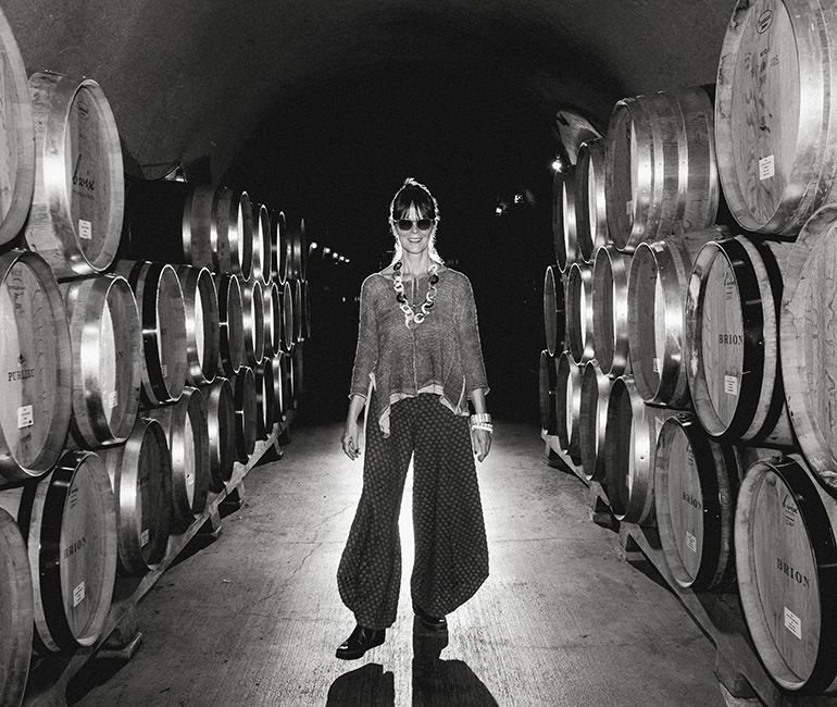 Person standing in a dim wine cellar aisle between rows of barrels, wearing sunglasses and wide-leg pants.
