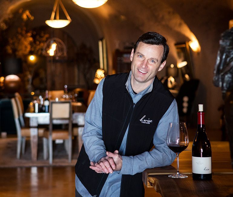 Man in a wine cellar stands beside a wine glass and bottle on a wooden table.