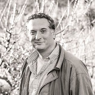 Man in a jacket and checkered shirt standing outdoors, smiling at the camera.