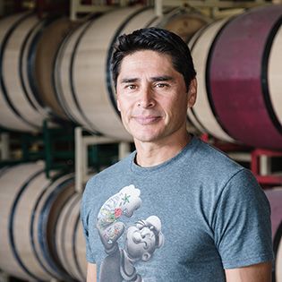 Man standing in a winery with stacked oak barrels, wearing a gray graphic T-shirt, slight smile