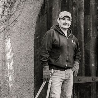 Man standing by a textured wall, holding two long tools, in a worn outdoor setting.