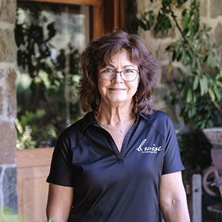 Woman in glasses and black polo shirt standing indoors with plants in the background
