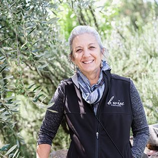 Smiling person in a black vest standing outdoors among olive trees.