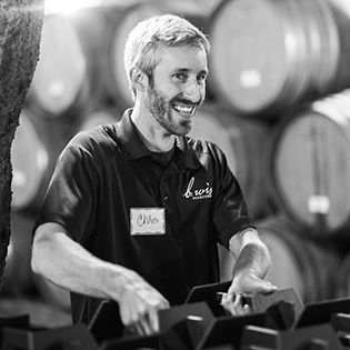 Smiling worker in a winery stands among stacked barrels, handling equipment.