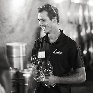 Smiling man in a black polo holding several wine glasses in a softly lit setting