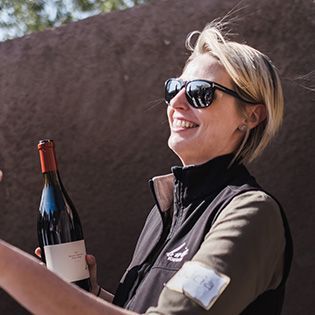 Smiling person in sunglasses holding a wine bottle outdoors.