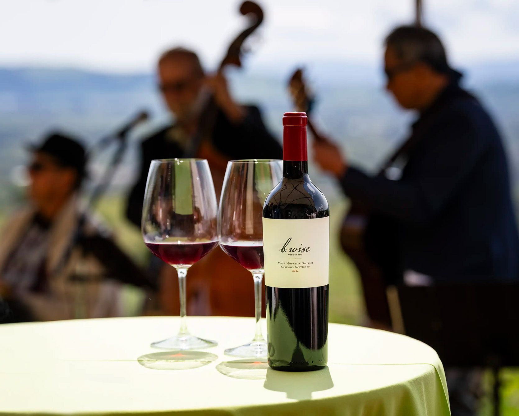 Wine bottle and two glasses on a table, with musicians playing blurred in the background outdoors.