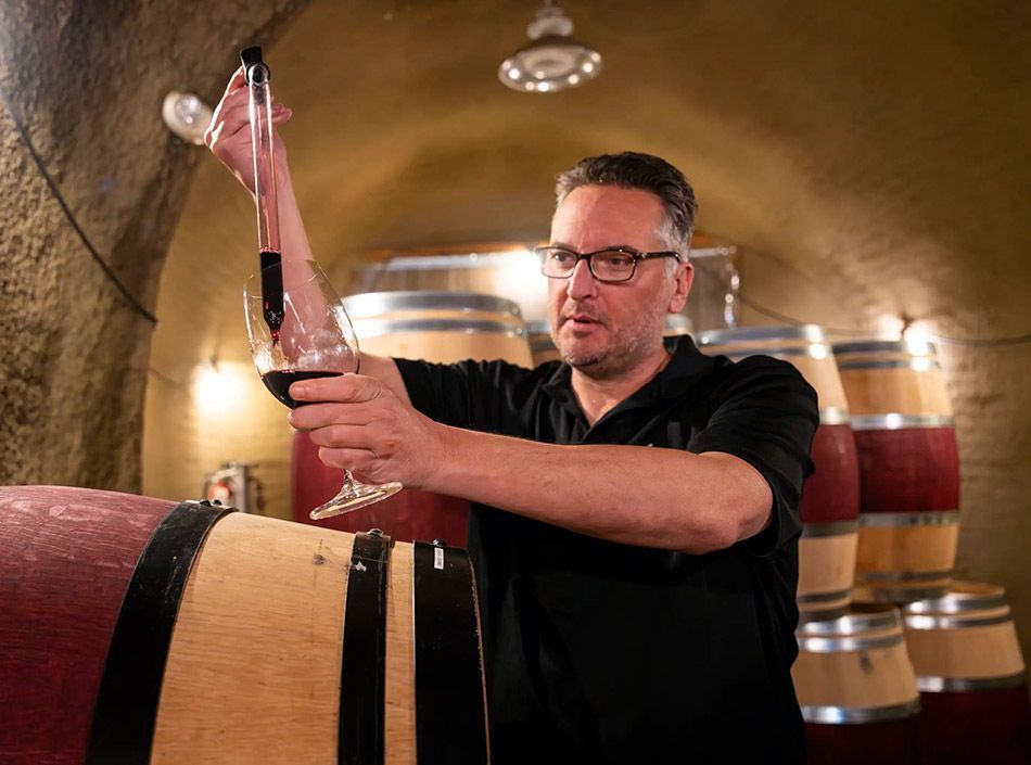Man in a wine cellar inspecting a bottle near wooden barrels under warm lighting