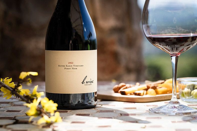 Wine bottle and glass beside bread on a sunlit outdoor table with yellow flowers