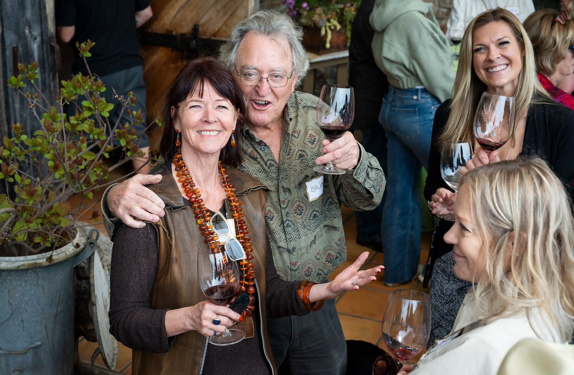 Four people chatting and smiling indoors, holding drinks in a cozy room with wood shelves and plants.