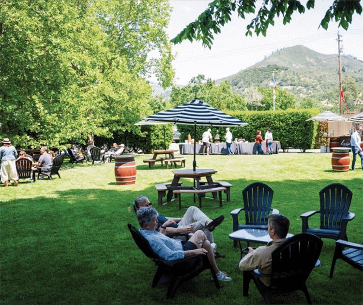 People relaxing on lawn chairs in a sunny park with tables, umbrellas, trees, and mountains in the background